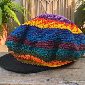 May include: A crocheted hat featuring a vibrant, multi-colored striped pattern in shades of purple, yellow, blue, green, and orange. The hat has a rounded, soft crown and a solid black, flat, circular brim extending from the front base.