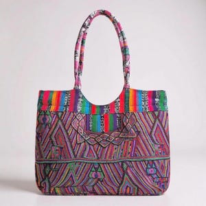 May include: A brightly coloured tote bag with a geometric pattern in red, purple, green, and blue. The bag has a rainbow striped top and matching handles. The design is eye-catching and unique, ideal for carrying essentials.