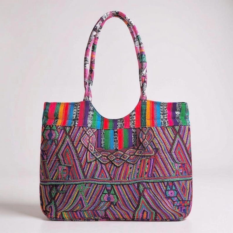 May include: A brightly coloured tote bag with a geometric pattern in red, purple, green, and blue. The bag has a rainbow striped top and matching handles. The design is eye-catching and unique, ideal for carrying essentials.