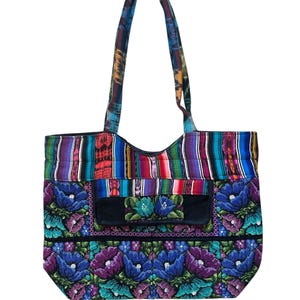 May include: A vibrant tote bag with a floral pattern. The bag has a black pocket with embroidered flowers and a striped top section. The handles are made of patterned fabric. The bag is approximately 38 cm wide and 36 cm tall.