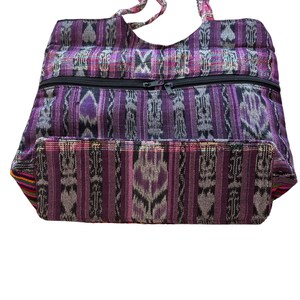 May include: A purple and grey patterned tote bag with a black zip closure. The bag has two handles and is made of a woven fabric. The bag's design features vertical stripes and geometric shapes.