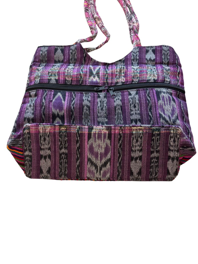 May include: A purple and grey patterned tote bag with a black zip closure. The bag has two handles and is made of a woven fabric. The bag's design features vertical stripes and geometric shapes.