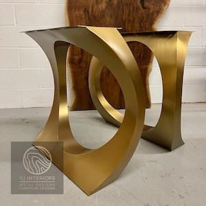 May include: Two gold-colored metal table bases with a unique, curved design. The bases have a rectangular top and a wide, open oval shape in the center. The logo "YJ INTERIORS METAL DESIGNS FURNITURE DESIGNS" is visible in the lower left corner.