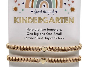 Pode incluir: Duas pulseiras de coração douradas com um fundo de arco-íris, estrelas e corações. O cartão diz "primeiro dia de KINDERGARTEN" e inclui uma mensagem sobre usar as pulseiras na escola.
