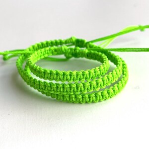 May include: Three neon green macrame bracelets are displayed against a white background. The bracelets are made of tightly knotted cord, with a textured appearance. The bracelets are adjustable and have long, loose ends.
