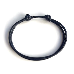 May include: A minimalist black bracelet with two knots. The bracelet is made of a thin, woven material and is designed to be worn around the wrist. The knots are located near the center of the bracelet.