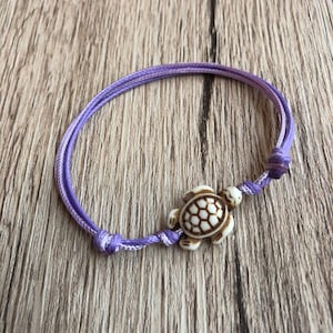 May include: A purple cord bracelet featuring a turtle charm. The turtle charm is off-white with a brown shell design. The bracelet is knotted at both ends and rests on a wooden surface.