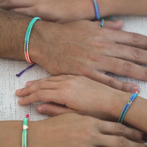 May include: A collection of four woven bracelets featuring vibrant neon color combinations of pink, blue, green, and purple. Each bracelet is crafted with a braided pattern and includes a small metallic heart-shaped charm on two of the pieces.