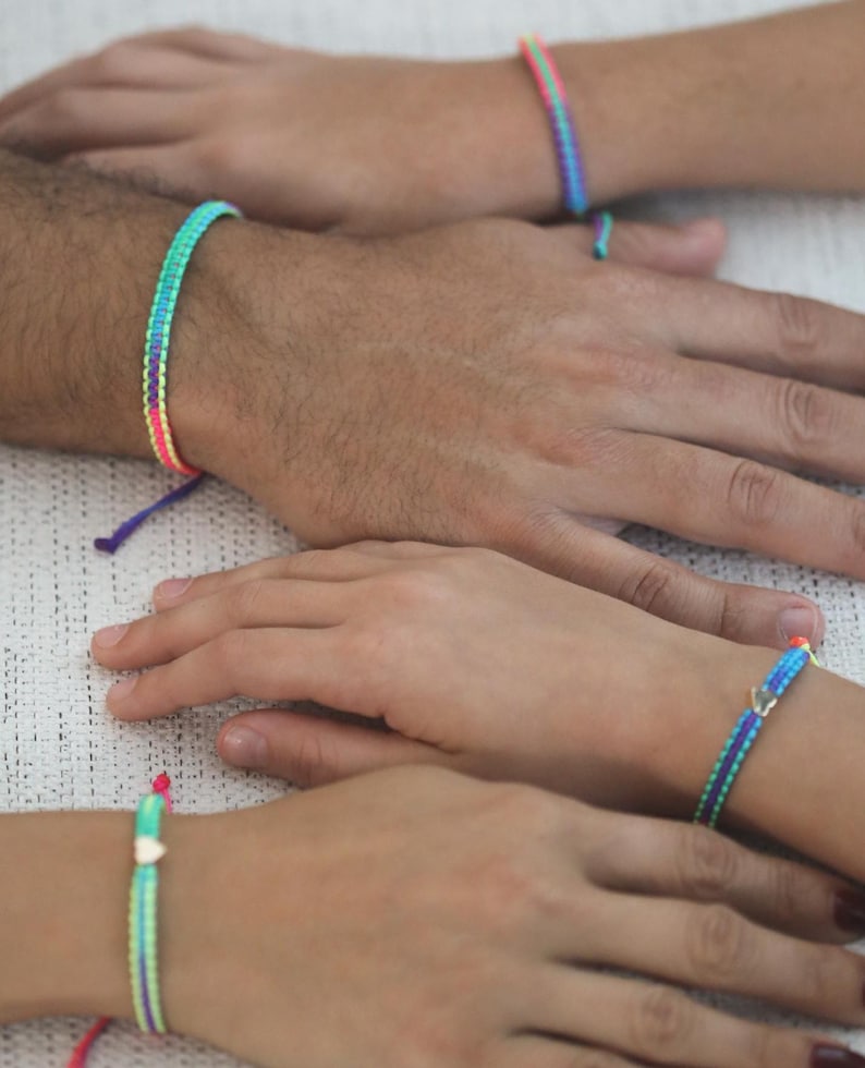May include: A collection of four woven bracelets featuring vibrant neon color combinations of pink, blue, green, and purple. Each bracelet is crafted with a braided pattern and includes a small metallic heart-shaped charm on two of the pieces.