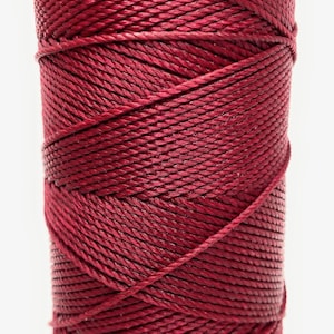 May include: A spool of deep red cord, wound tightly in a cylindrical shape. The cord is smooth and appears to be made of a synthetic material.