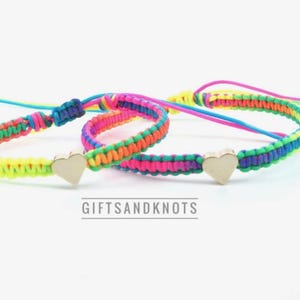 May include: Two woven cord bracelets featuring neon yellow, pink, blue, and green threads with a central metallic heart charm on each. The bracelets have adjustable sliding knots and the text GIFTSANDKNOTS is printed below the items.
