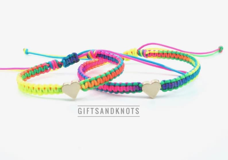 May include: Two woven cord bracelets featuring neon yellow, pink, blue, and green threads with a central metallic heart charm on each. The bracelets have adjustable sliding knots and the text GIFTSANDKNOTS is printed below the items.