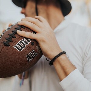 May include: A brown leather braided bracelet with a black clasp. The bracelet is worn on the wrist of a person holding a brown football with white laces and the text "Wilson All American" printed on the side.