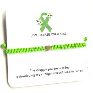 May include: A neon green macrame bracelet with a small heart charm. The bracelet is displayed on a white card with the text "LYME DISEASE AWARENESS" and a green ribbon graphic. The card also includes the quote: "The struggle you are in today is developing the strength you will need tomorrow."