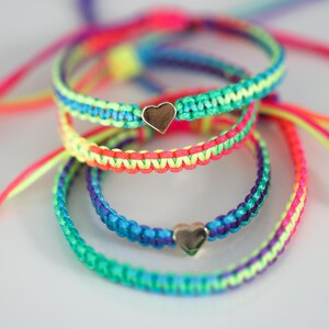May include: A collection of four braided cord bracelets in vibrant neon colors including pink, yellow, blue, purple, and green. Two of the bracelets feature a small, polished metallic heart-shaped charm centered on the woven cord band.
