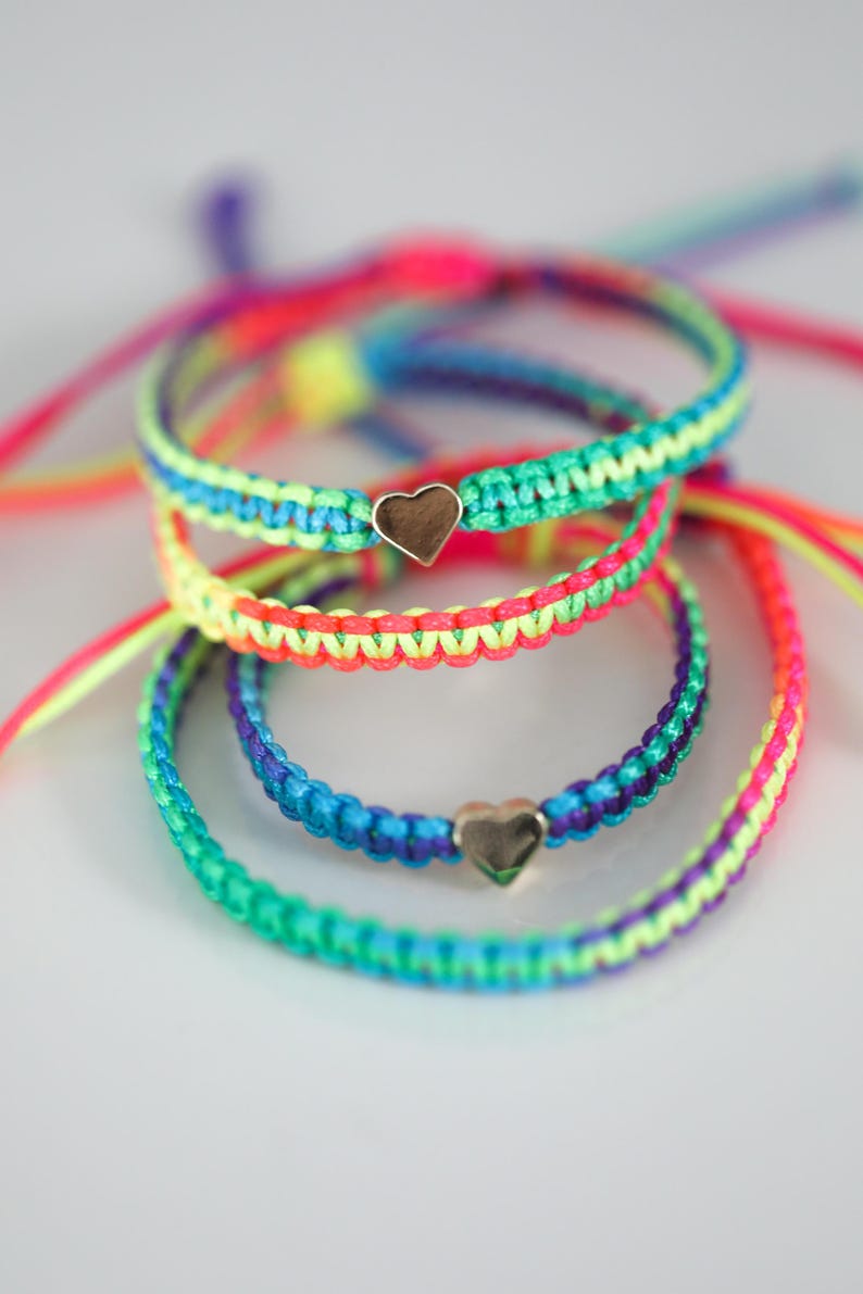 May include: A collection of four braided cord bracelets in vibrant neon colors including pink, yellow, blue, purple, and green. Two of the bracelets feature a small, polished metallic heart-shaped charm centered on the woven cord band.
