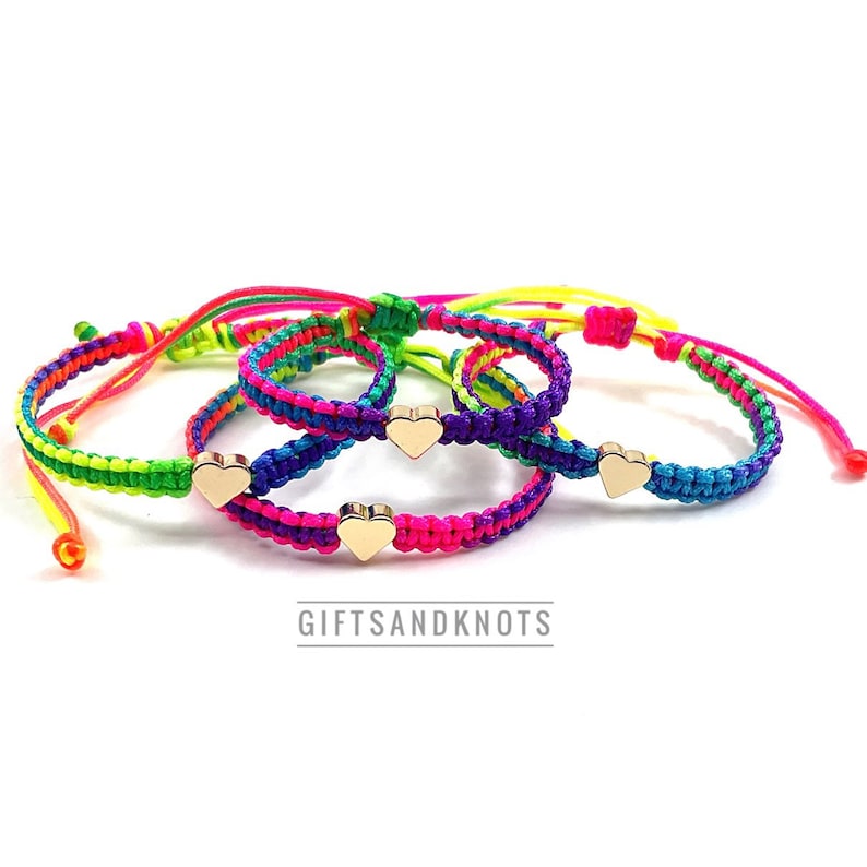 May include: Three adjustable bracelets made of neon-colored braided cord in shades of pink, green, blue, and purple. Each bracelet features a small, polished gold-tone metal heart charm at the center. The text GIFTSANDKNOTS is printed below the items.