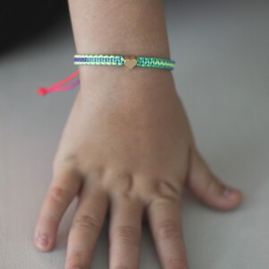 May include: A woven cord bracelet featuring a central gold-toned heart charm. The band is constructed from alternating light green, purple, and teal threads, finished with bright pink adjustable pull strings at the end for a secure fit.