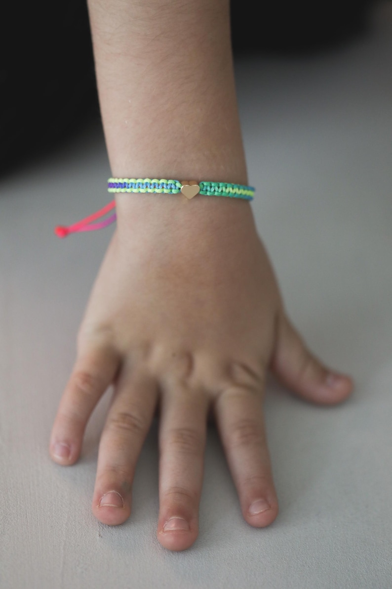 May include: A woven cord bracelet featuring a central gold-toned heart charm. The band is constructed from alternating light green, purple, and teal threads, finished with bright pink adjustable pull strings at the end for a secure fit.