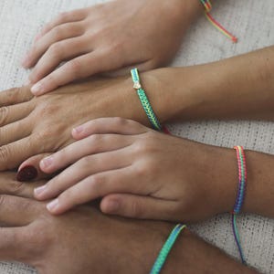May include: Four woven bracelets featuring vibrant neon rainbow patterns. Each bracelet is constructed with intricate knotting techniques, displaying bright color gradients of pink, yellow, green, and blue. One bracelet includes a small metallic heart charm.