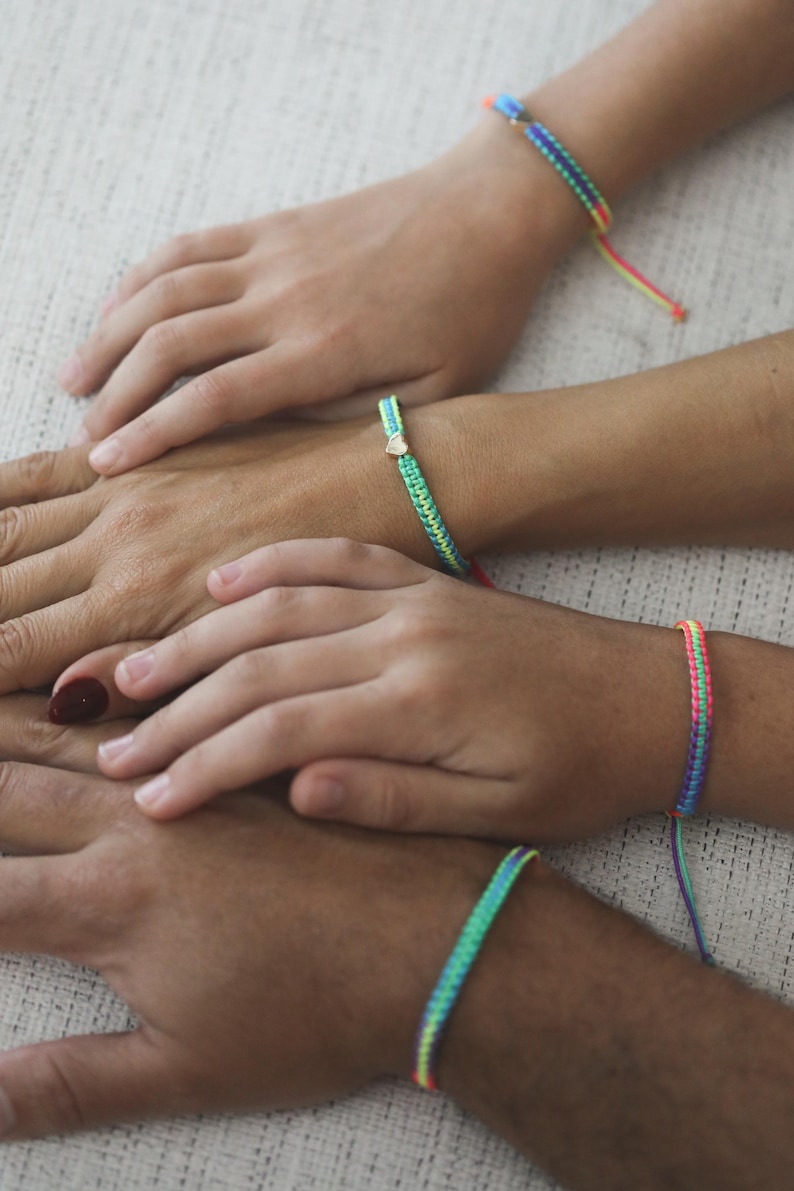 May include: Four woven bracelets featuring vibrant neon rainbow patterns. Each bracelet is constructed with intricate knotting techniques, displaying bright color gradients of pink, yellow, green, and blue. One bracelet includes a small metallic heart charm.