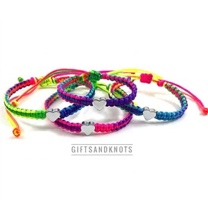 May include: Three braided bracelets featuring neon pink, green, blue, and purple cord patterns. Each bracelet includes a small, polished silver-tone metal heart charm centered on the band. The text GIFTSANDKNOTS is printed below the bracelets.