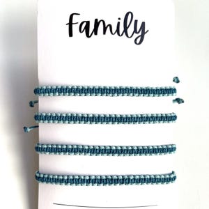 May include: Four blue and light blue woven bracelets are displayed on a white card. The word "Family" is printed in black script at the top of the card. The bracelets are made of a woven material.