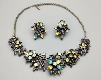 Op de afbeelding: Een zilverkleurige ketting en oorbellenset met een bloemmotief. De ketting heeft een grote bloem met blauwe, gele en lichtblauwe strasssteentjes. De oorbellen zijn kleiner en hebben een vergelijkbaar bloemmotief met blauwe en gele strasssteentjes.