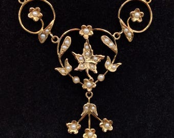 May include: A gold-colored necklace with a floral design. The necklace features small pearls and flower-shaped accents. The central pendant has a star-shaped design with pearls. The necklace is on a black background.