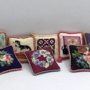 Pode incluir: Oito pequenos travesseiros quadrados de ponto de agulha com vários desenhos. Os travesseiros são em tons de verde, azul, branco, vermelho, preto e roxo. Os desenhos incluem flores, um cão, um padrão de colcha e um gato.