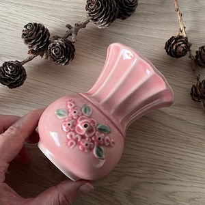 Puede incluir: Un jarrón de cerámica rosa con un diseño floral. El jarrón tiene una boca ancha y una base estrecha. Está decorado con un grupo de flores rosas y hojas verdes.