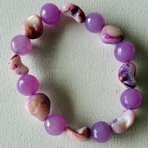 May include: A bracelet featuring alternating round purple beads and irregularly shaped, light pink and brown shell beads. The bracelet is displayed on a light-colored, textured surface.