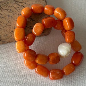 May include: A bracelet composed of smooth, rectangular orange beads with a glossy finish. The strand features a single, irregular white pearl accent, creating a contrast against the vibrant orange tones of the uniform, polished stone beads.
