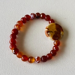 May include: A red carnelian gemstone bracelet with a large, round, white and brown agate charm.
