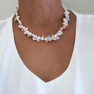 May include: A necklace composed of irregularly shaped, off-white pearls. The pearls are strung together, creating a unique and organic design. The necklace is worn against a white top, highlighting the natural color of the pearls.