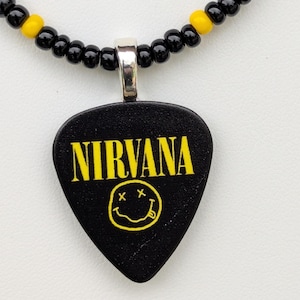 May include: Black guitar pick pendant necklace featuring the word "NIRVANA" in yellow, with a yellow smiley face. The necklace has a black beaded chain with yellow accents.