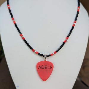 May include: A necklace featuring a red guitar pick pendant with the name ADELE printed in black capital letters. The pendant hangs from a beaded chain composed of small black, red, and silver-toned beads arranged in a repeating pattern.