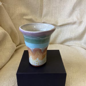 May include: A ceramic vase with a wide mouth and a flared base. The vase is decorated with a multi-colored design of purple, green, blue, orange, and yellow.