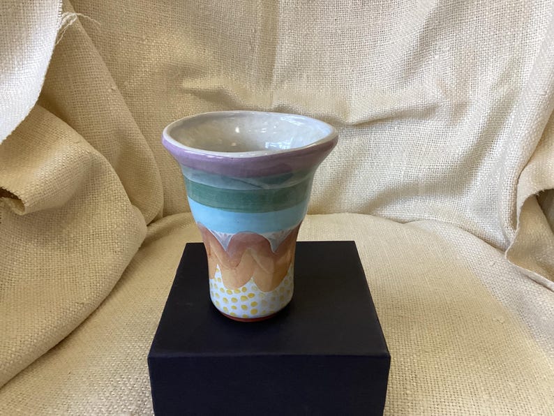 May include: A ceramic vase with a wide mouth and a flared base. The vase is decorated with a multi-colored design of purple, green, blue, orange, and yellow.