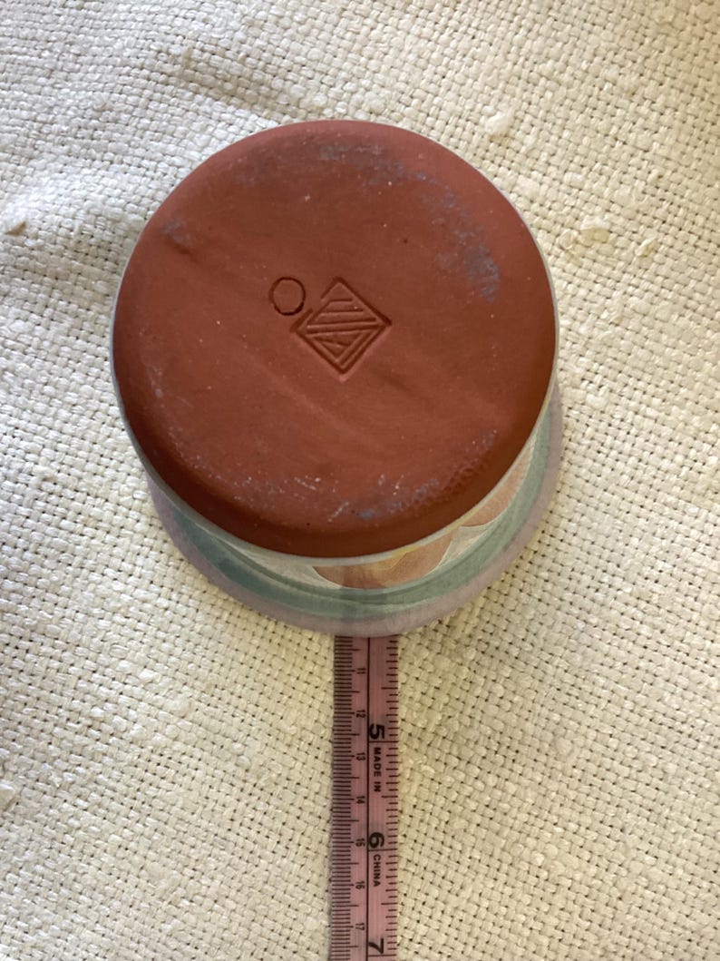 May include: A terracotta planter with a light blue, green, and orange design. The bottom of the planter has a stamped mark with a circle and a diamond shape.