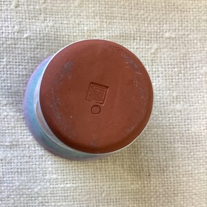 May include: The bottom of a ceramic cup with a light blue, pink, and white glaze. The bottom of the cup is brown and has a square mark with a circle inside.