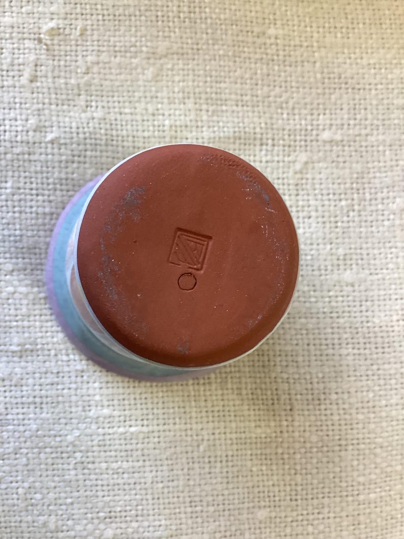 May include: The bottom of a ceramic cup with a light blue, pink, and white glaze. The bottom of the cup is brown and has a square mark with a circle inside.