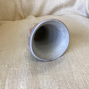 May include: A white ceramic vase with a purple rim and a light brown, yellow, and white floral design. The vase is sitting on a beige fabric surface.