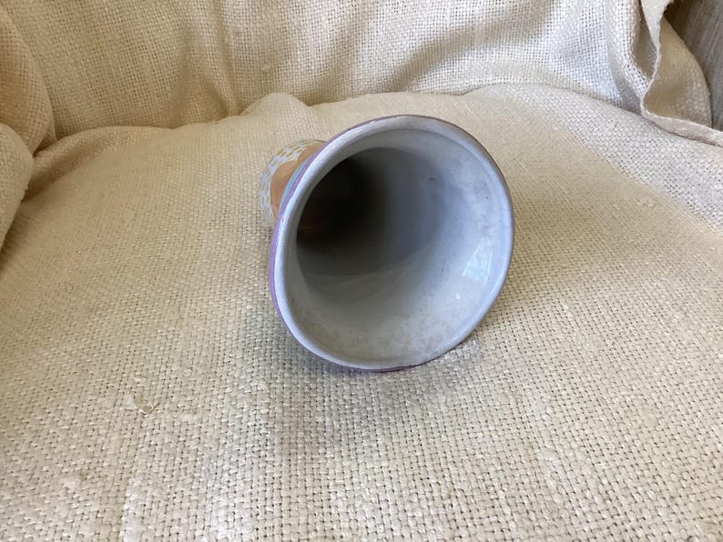 May include: A white ceramic vase with a purple rim and a light brown, yellow, and white floral design. The vase is sitting on a beige fabric surface.