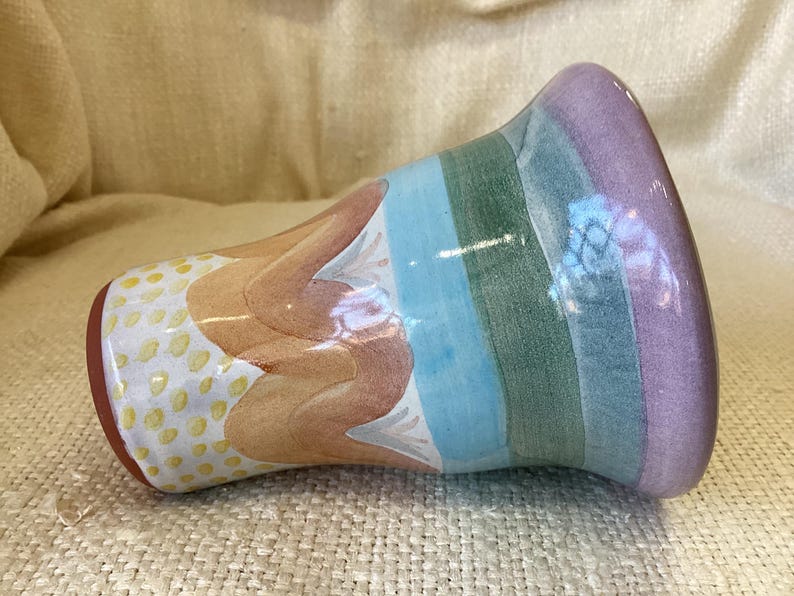 May include: A colorful ceramic vase with a striped design in shades of purple, blue, green, and orange. The vase has a textured, speckled pattern on the bottom.
