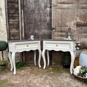 May include: Two white painted side tables with a single drawer and curved legs. The tables have a grey top and gold drawer pulls.