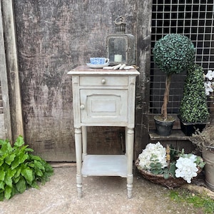May include: A vintage-style, light grey bedside table with a drawer and a cabinet door. A blue and white teacup and saucer are on the tabletop, alongside a decorative lantern. The table has a lower shelf and stands on four turned legs.