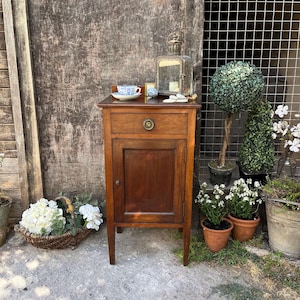 May include: A wooden bedside table with a single drawer featuring a round metal pull and a lower cabinet door. The top surface holds a blue and white patterned teacup on a saucer, a small decorative object, and a metal lantern with a glass pane.