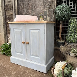 May include: A light blue cabinet with two doors and wooden knobs. The cabinet has a wooden top and a base with a decorative moulding. The cabinet is sitting on a concrete floor.