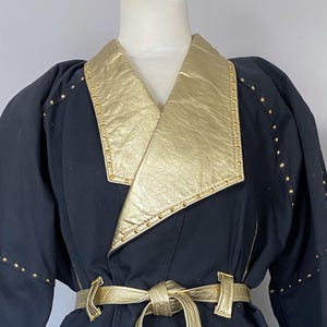 May include: Black skirt suit with gold accents. The jacket has a gold collar and cuffs, and the skirt is knee-length. Gold studs adorn the jacket, skirt, and cuffs. A gold belt cinches the waist, creating a vintage, stylish look.